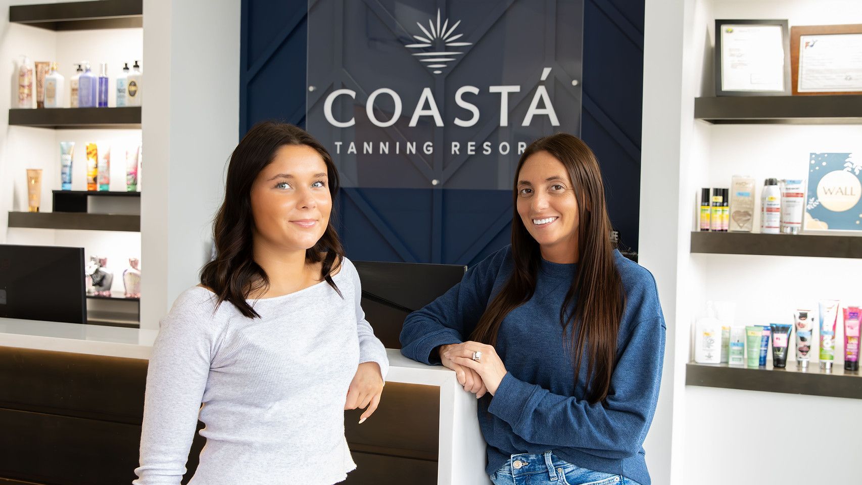 Two women stand at a tanning salon reception desk with COASTA branding and product shelves behind them