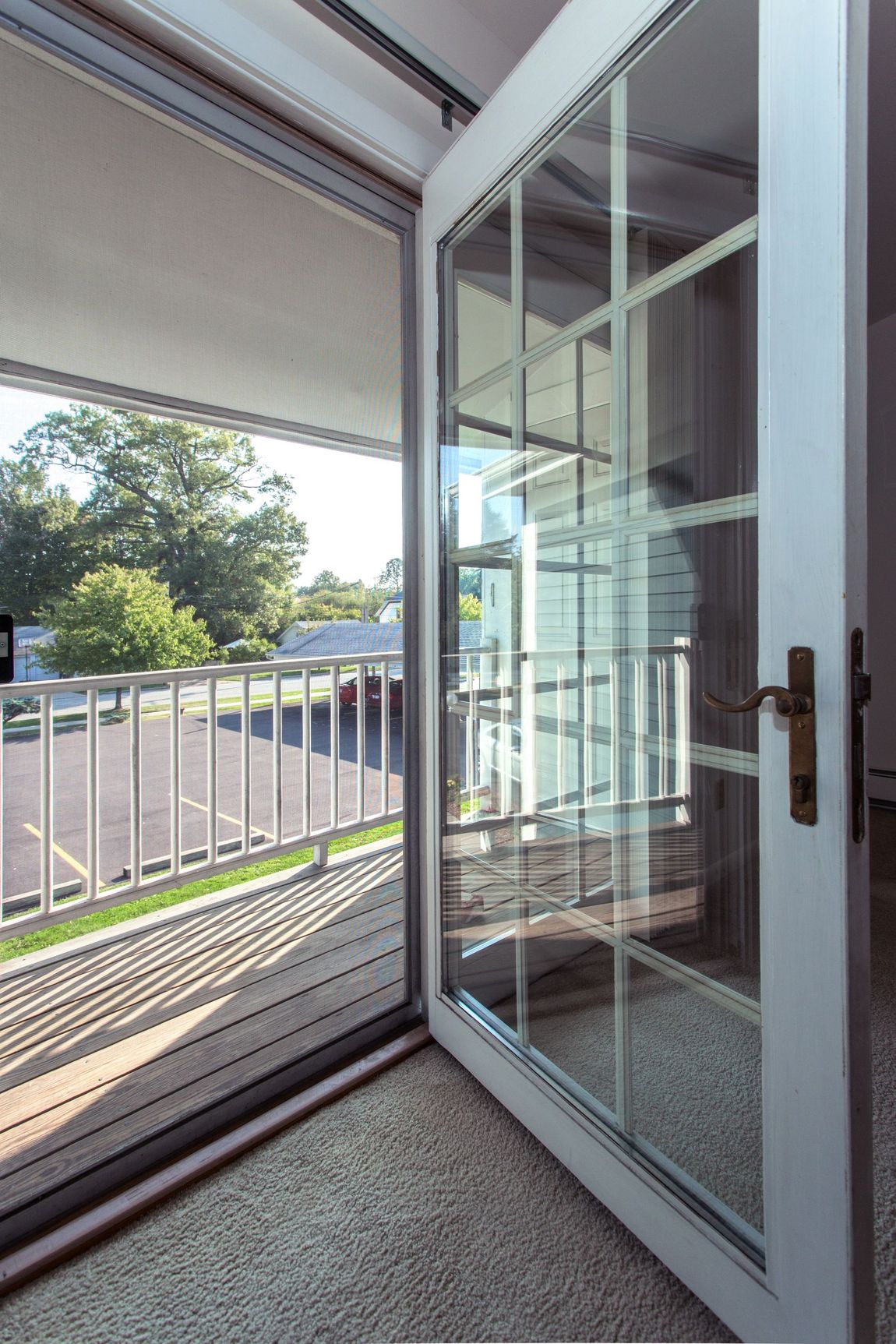 Open white French door with screen leading to a balcony with a white railing. Trees and parking lot visible outside.