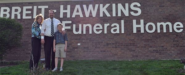Three people stand outside the Breit-Hawkins Funeral Home, which has large white lettering on a brick wall.