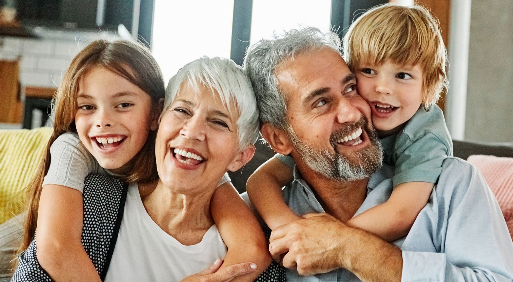 A smiling girl and boy embrace an older couple in a brightly lit indoor space, all looking happy.