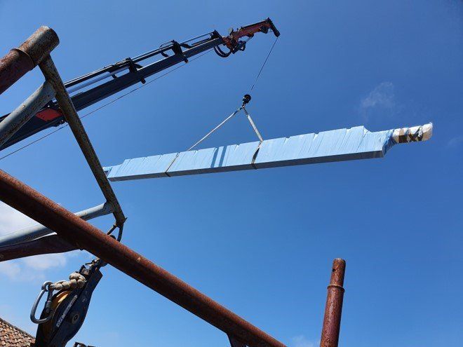 Un lungo oggetto ricoperto di blu, forse un pannello del tetto, viene sollevato da una gru contro un cielo azzurro e terso.