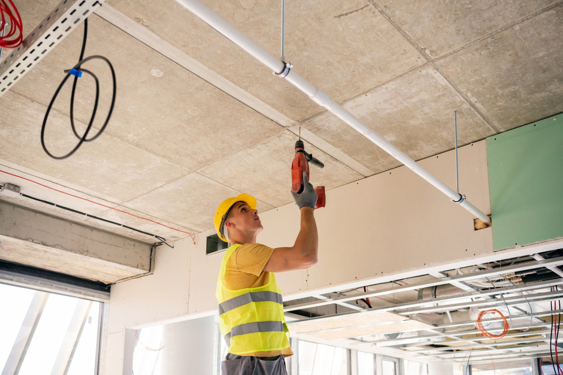 Un operaio edile con un casco giallo usa un trapano su un soffitto con tubi e cavi.