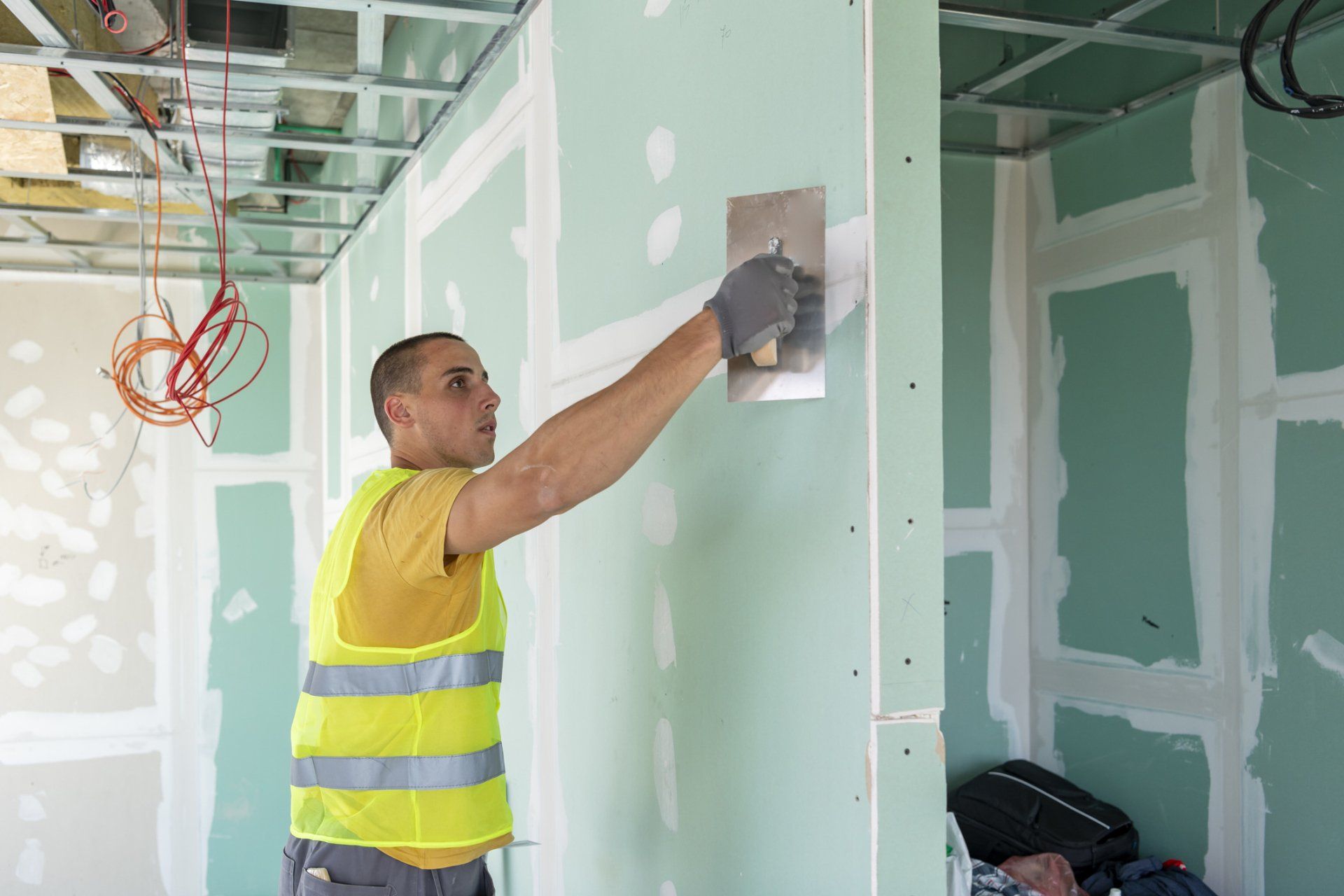 Uomo con giubbotto di sicurezza che applica il composto per cartongesso con una cazzuola su un muro parzialmente costruito.