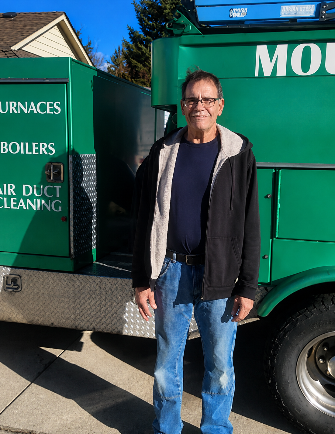 Man standing beside a green service truck with furnace and boiler lettering, outdoors in sunlight