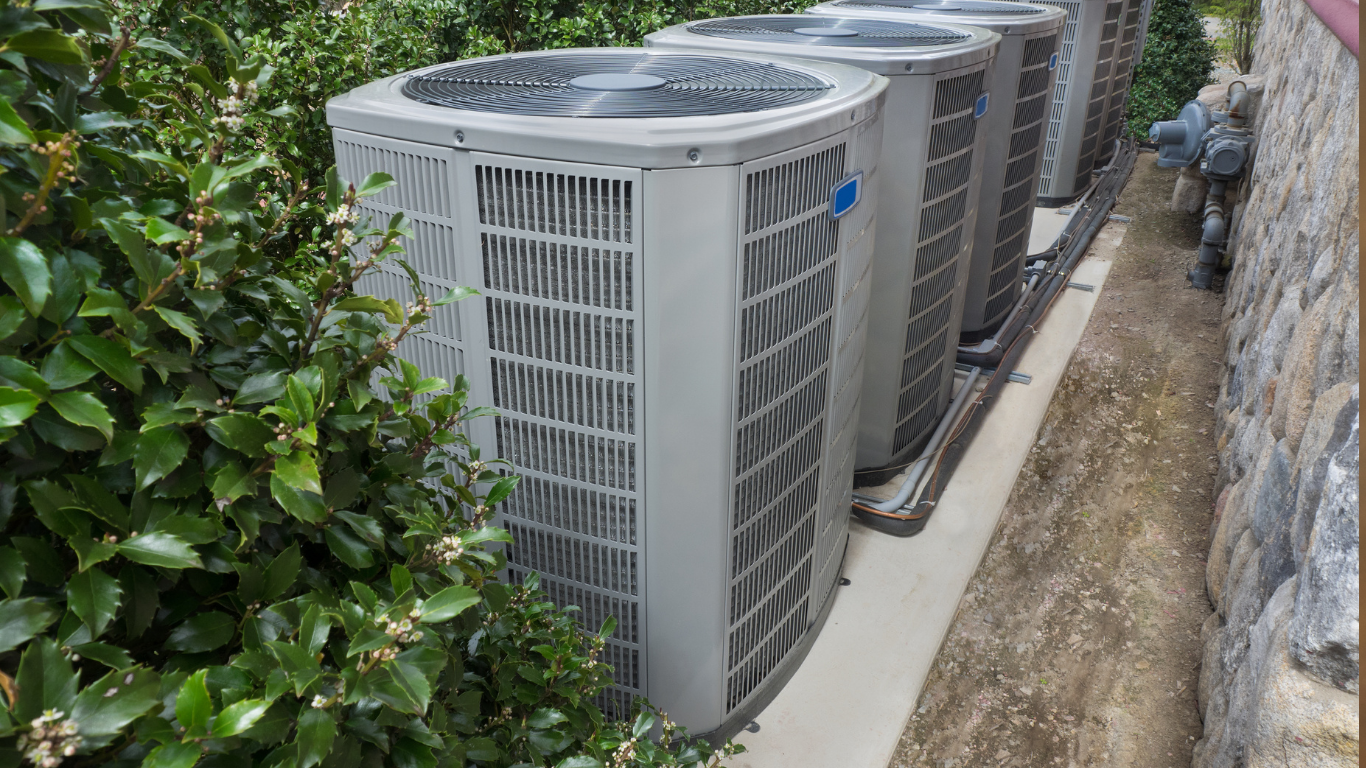 Row of outdoor air conditioning units beside a building and stone wall, with bushes on the left.