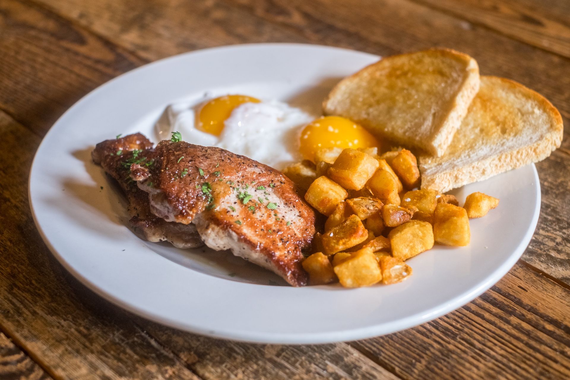 A white plate topped with meat , eggs , potatoes and toast on a wooden table.