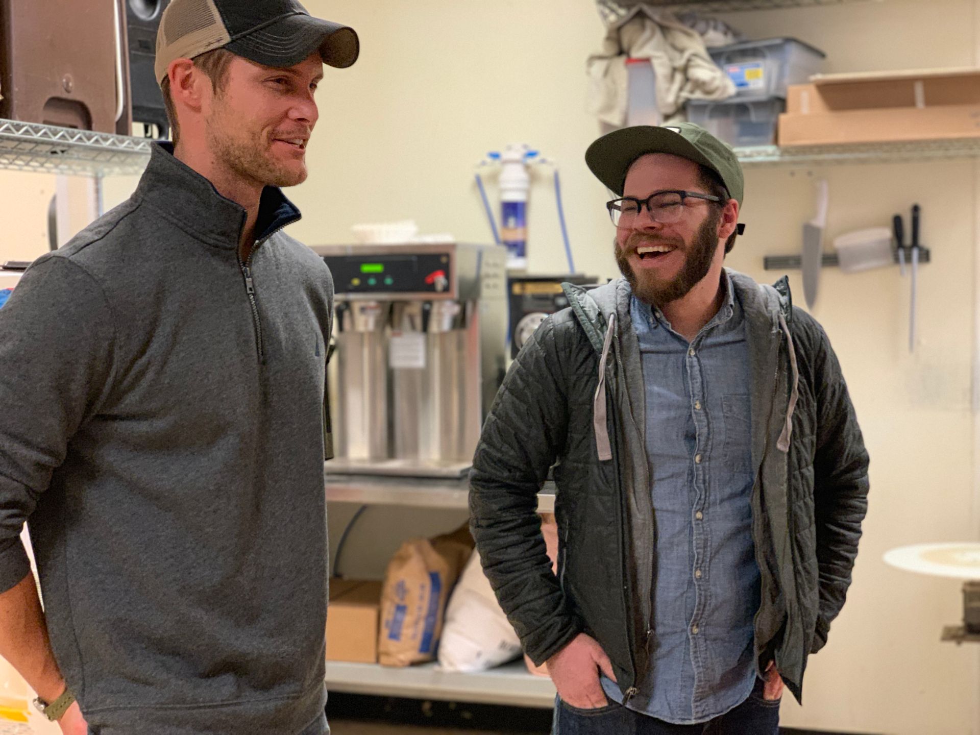 Two men are standing next to each other in a kitchen and smiling.