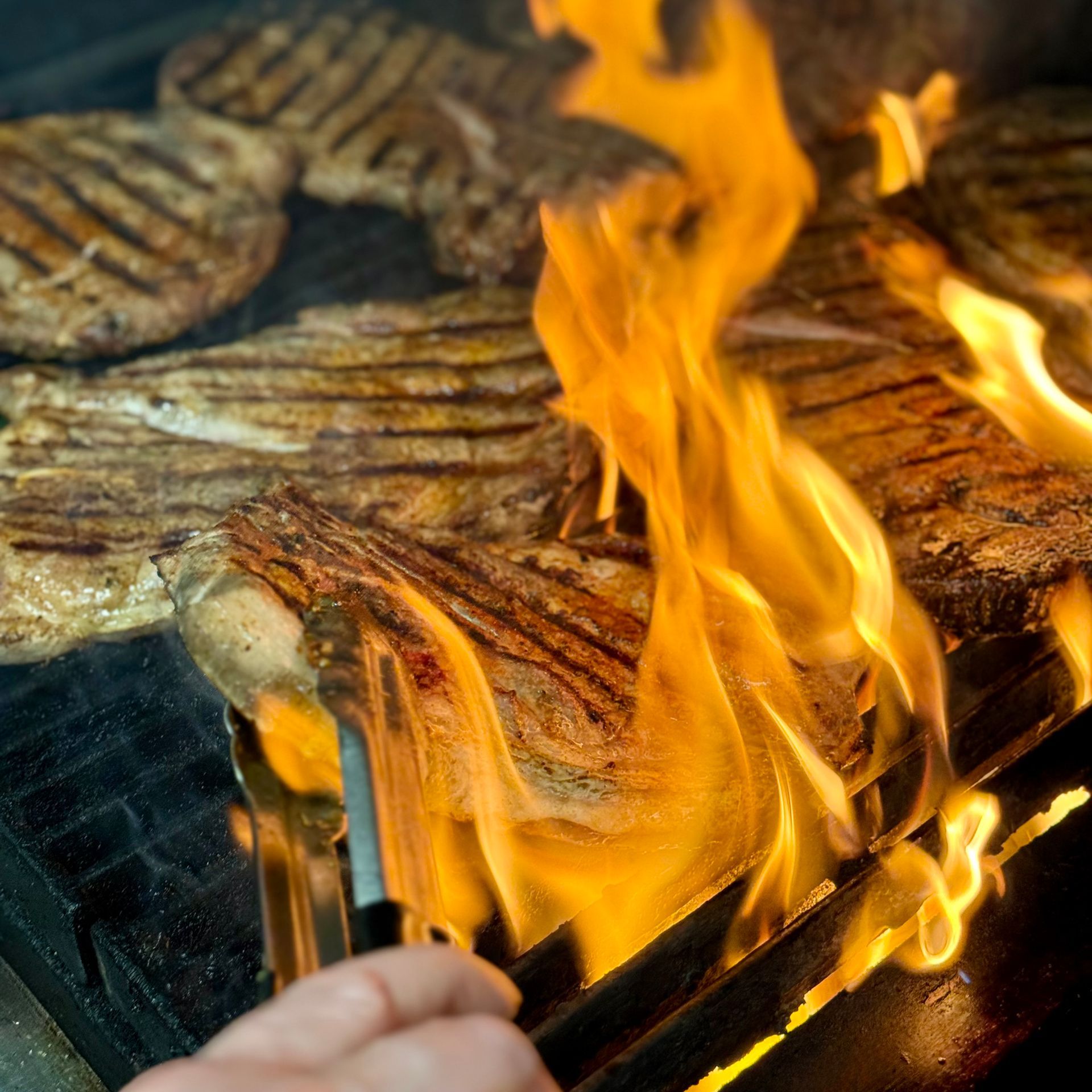 A person is holding a pair of tongs over a grill with flames coming out of it