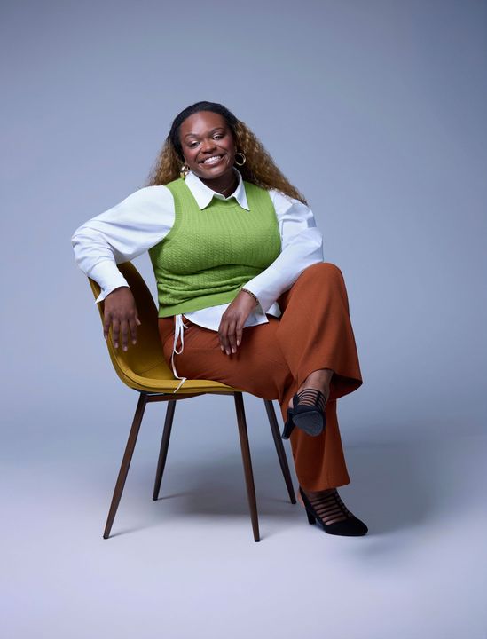 Woman seated, smiling. She wears a white blouse, green vest, rust-colored pants, and black shoes, leaning on a yellow chair.