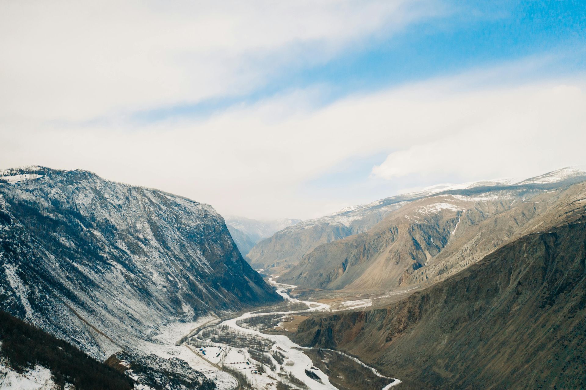 Snowy mountain valley with a winding river under a cloudy blue sky.