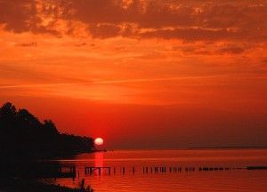 Sunset over water with a bright orange sky. Silhouette of land on the left, posts in the water.