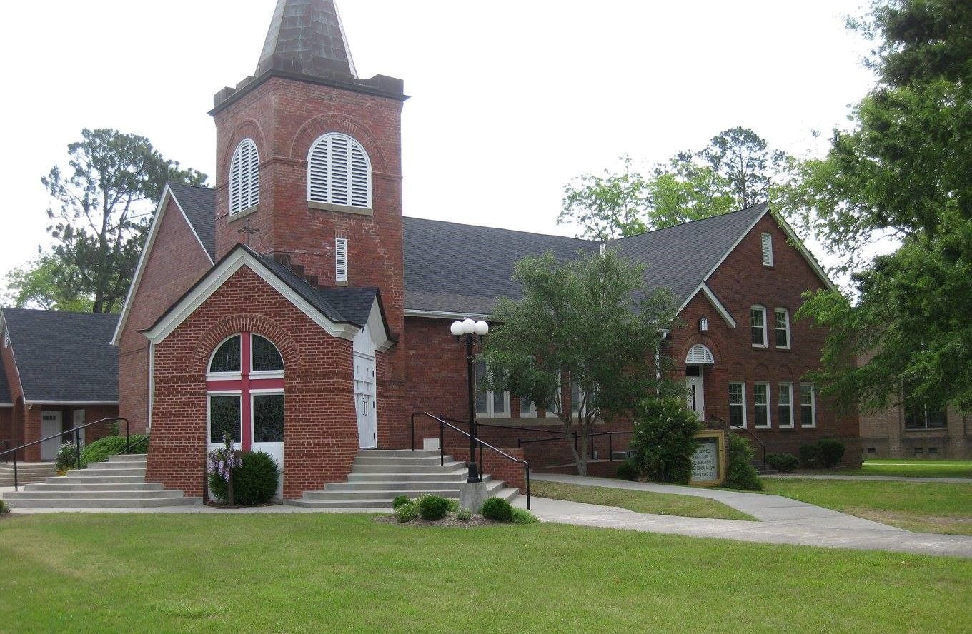 Brick church building with a steeple, white arched windows, and a grassy lawn.