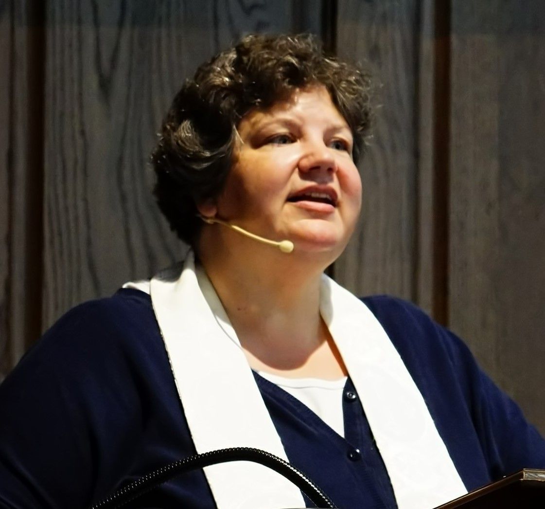 Pastor speaking at a lectern, wearing a dark blue sweater and white collar; microphone near her mouth.