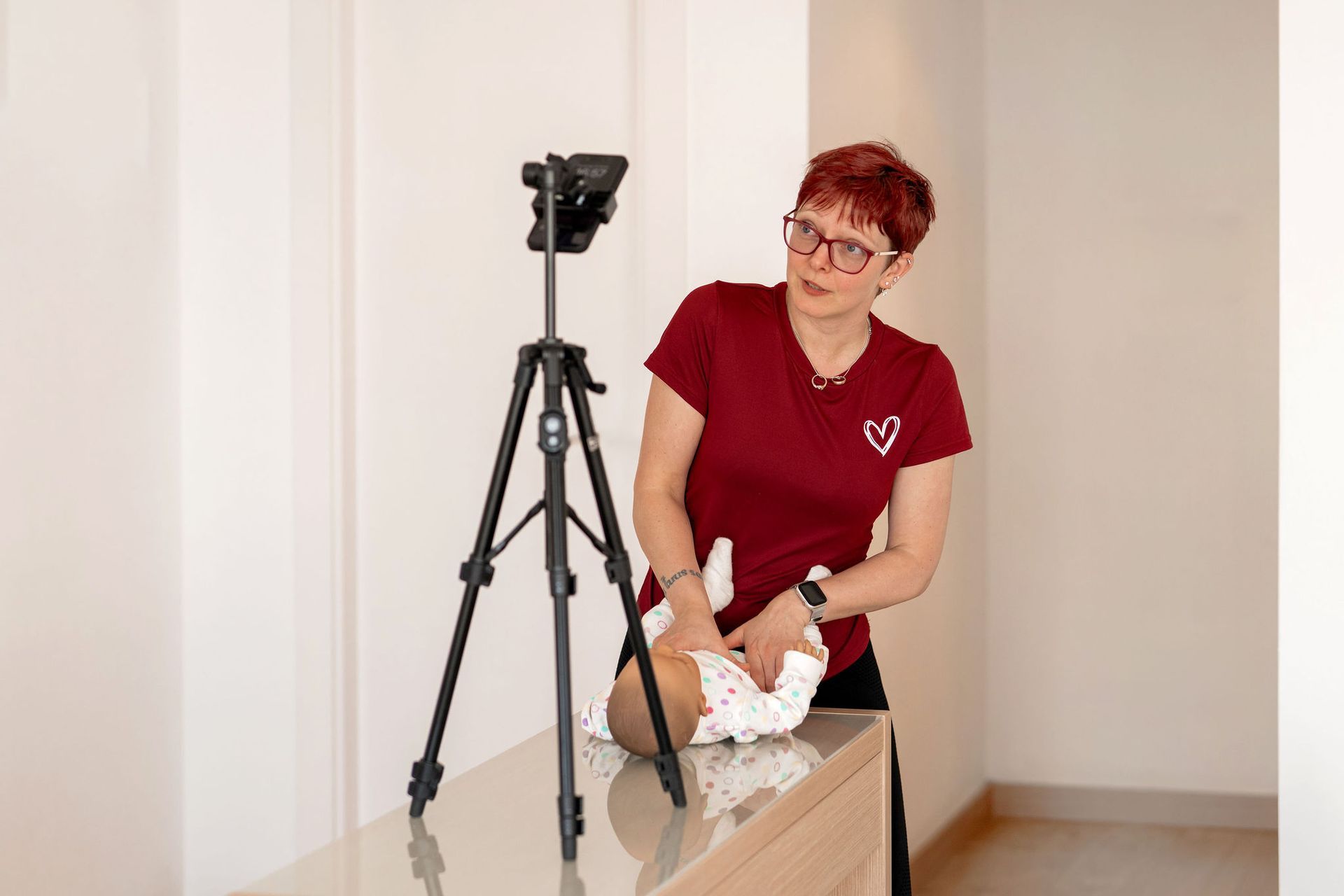 Eine Frau mit kurzen roten Haaren und Brille demonstriert die Verwendung einer Babypuppe, Stativ mit montierter Kamera.