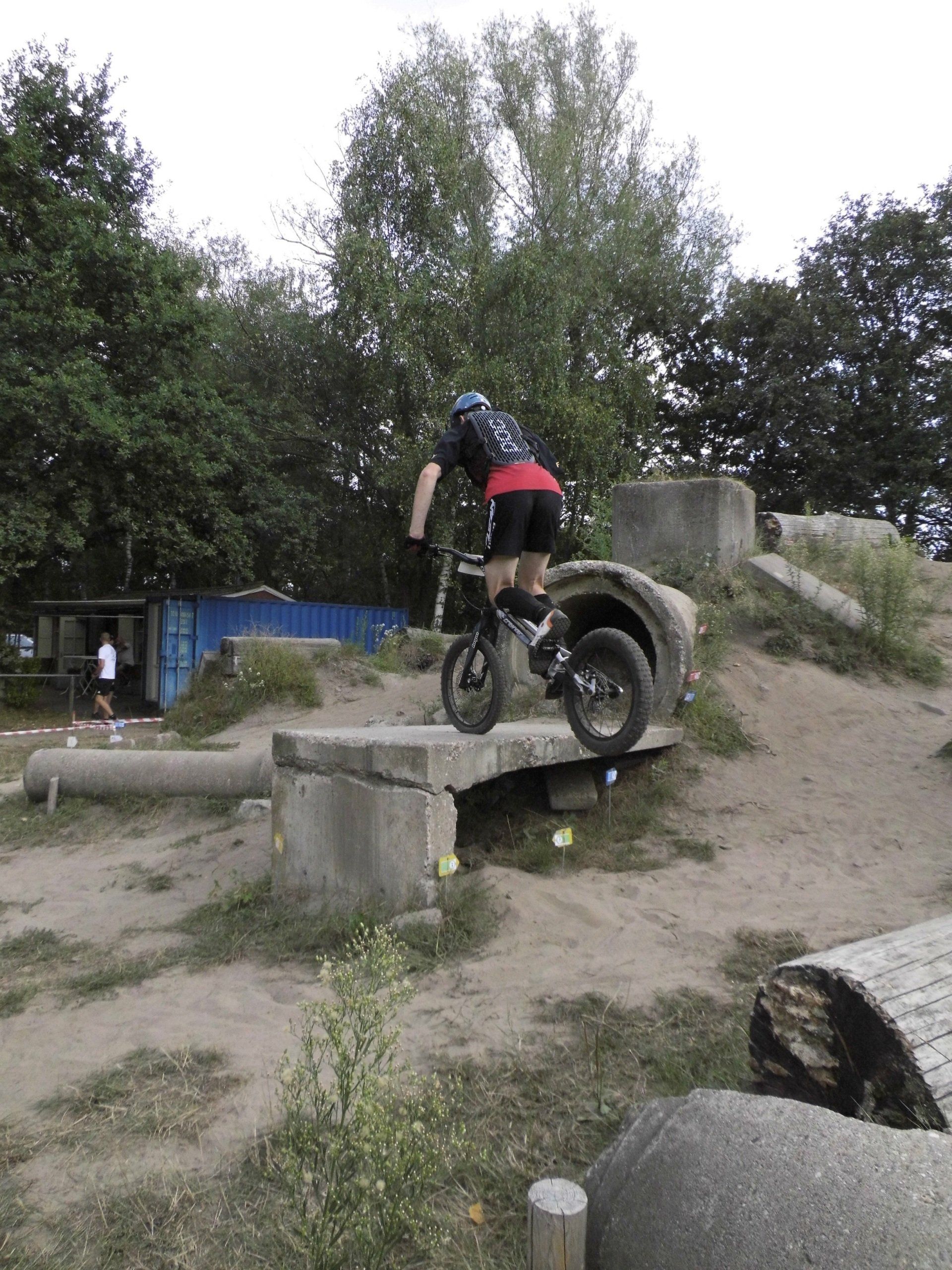 Een mountainbiker rijdt over een betonnen obstakel op een onverhard pad, met bomen en rotsen op de achtergrond.