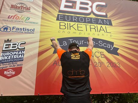 Een persoon met oranje mouwen heft zijn armen op voor een spandoek van het EBC European Bikers Trial-evenement.