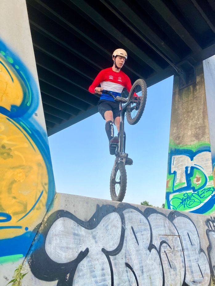 Een fietser maakt een sprong in de lucht onder een brug, gekleed in een rood shirt en helm, met graffiti op de muren eronder.