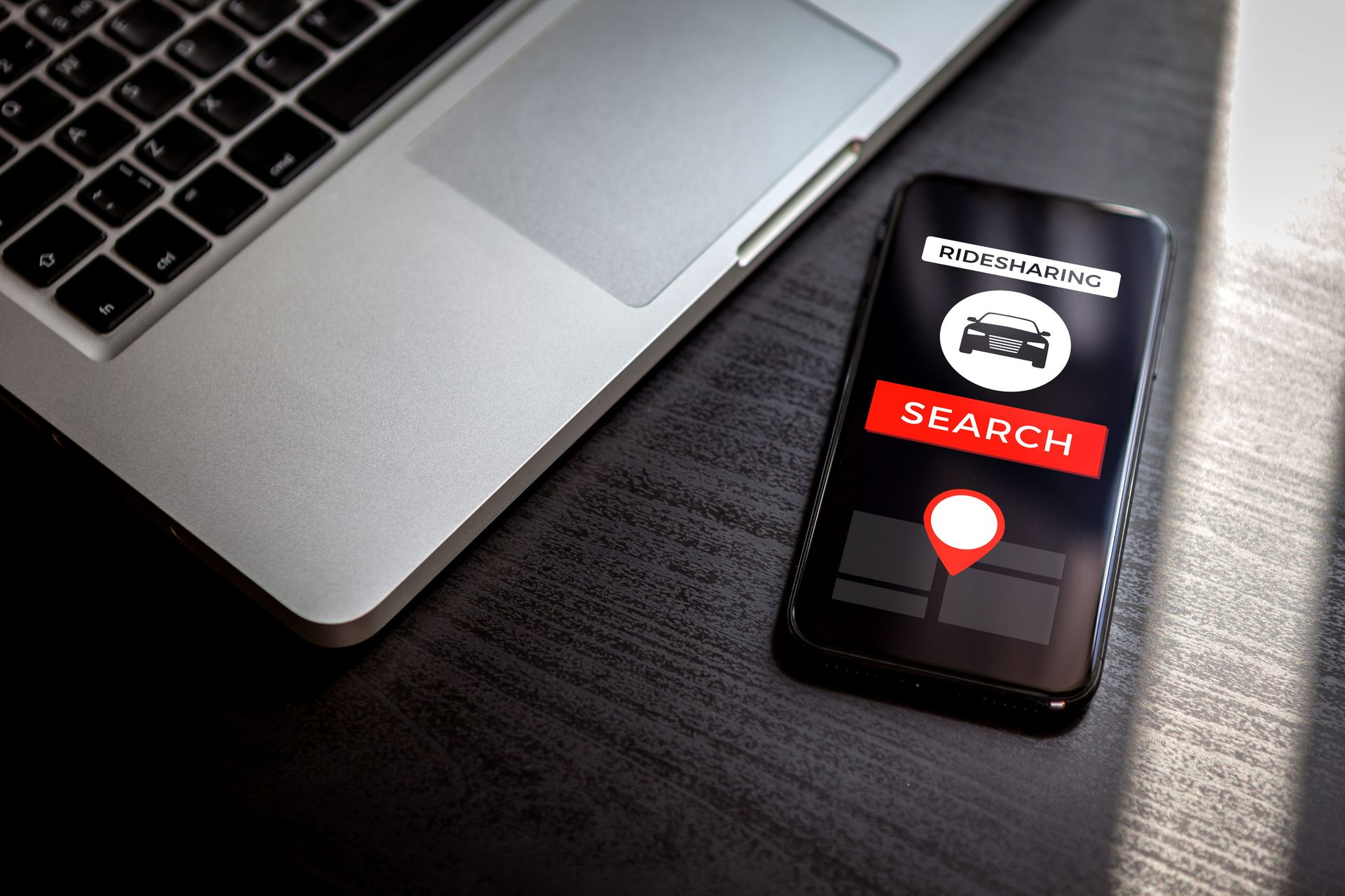 Laptop beside smartphone showing a “SEARCH” screen with a red map pin on a dark desk.