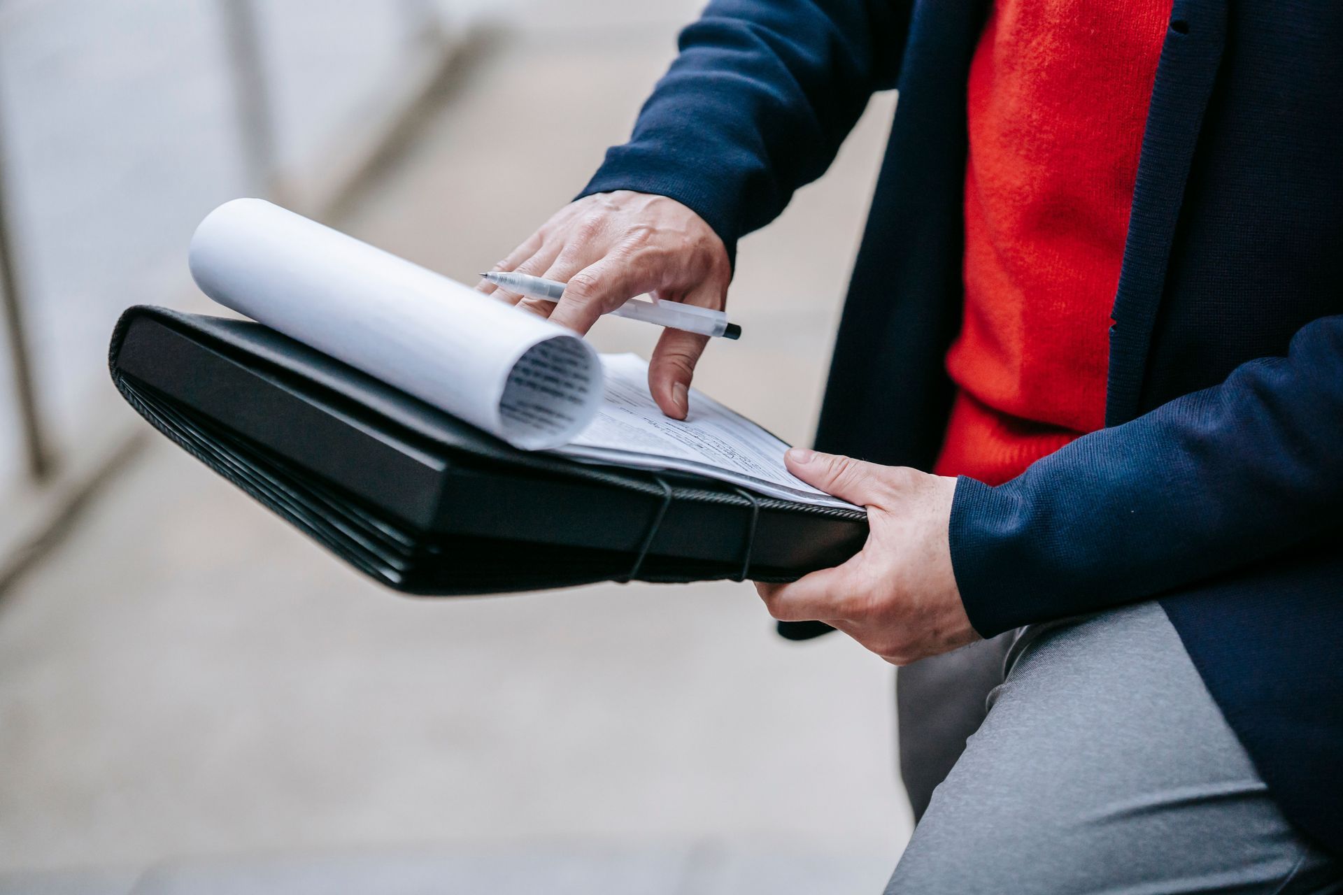 Person in a dark suit and red tie holds an open folder and papers outdoors.