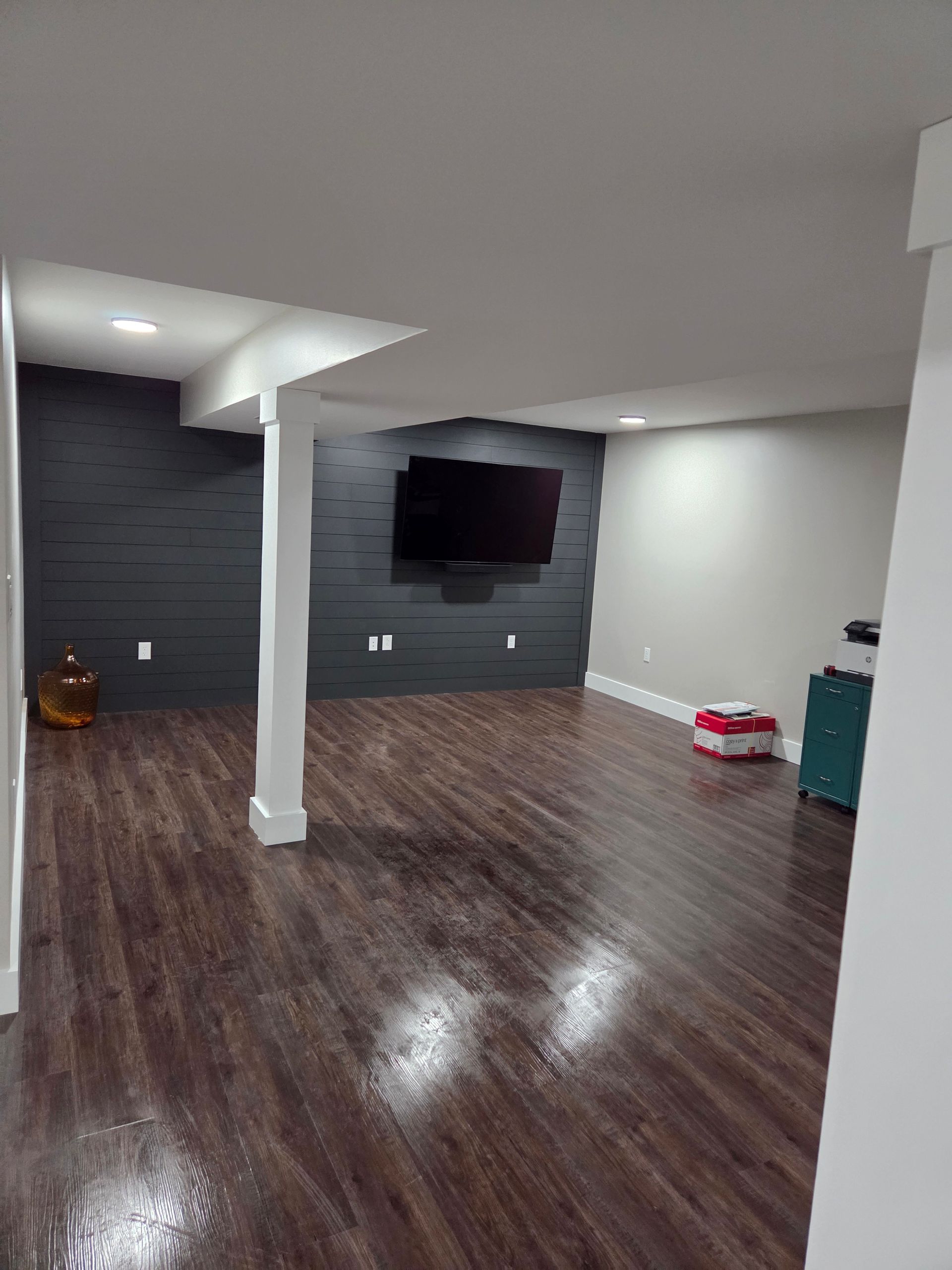 A modern basement with dark wood floors, a shiplap accent wall, a wall-mounted TV, and a white support column.