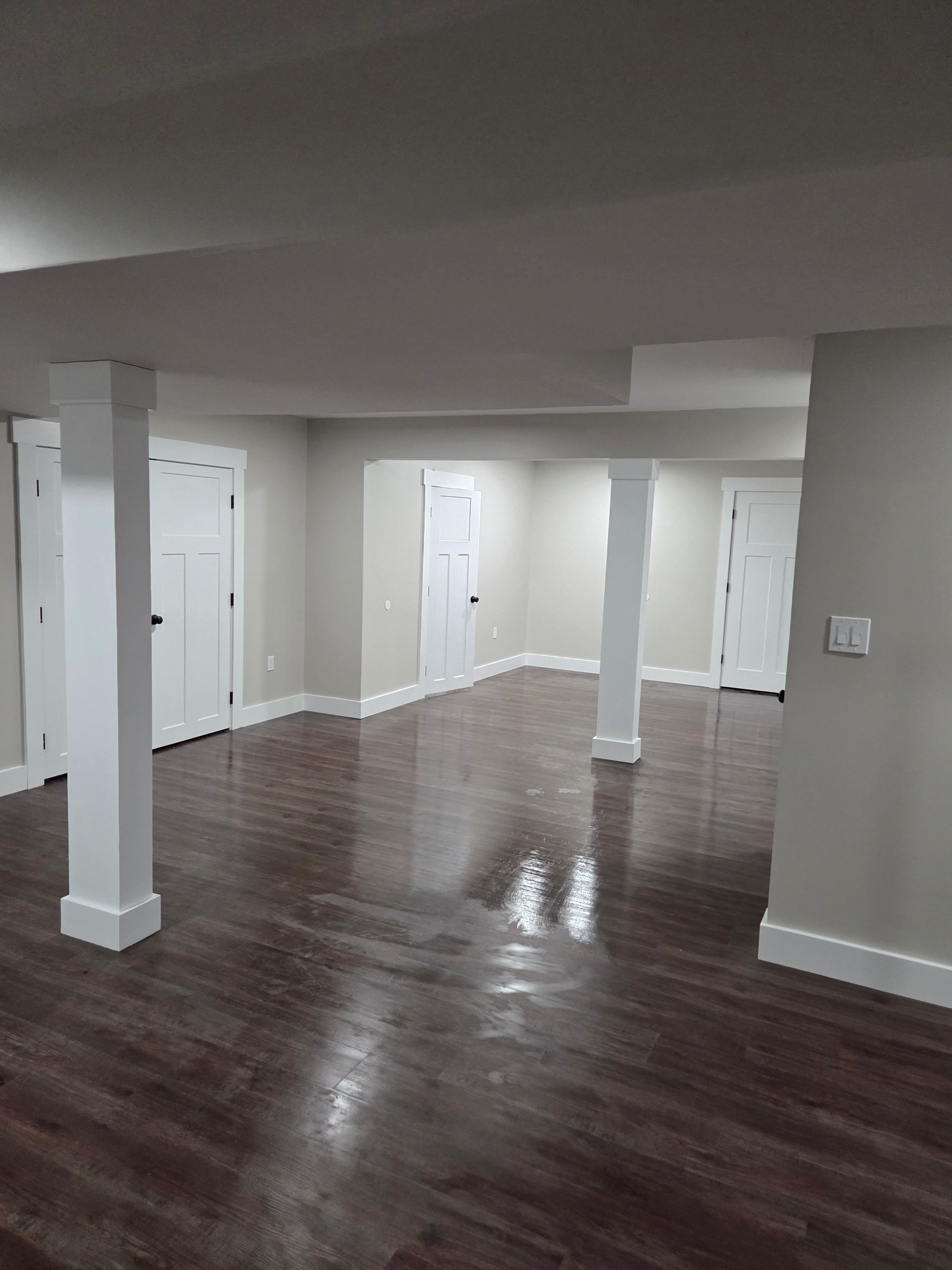 A finished basement with wood-look flooring, light grey walls, white trim, and two square support columns.
