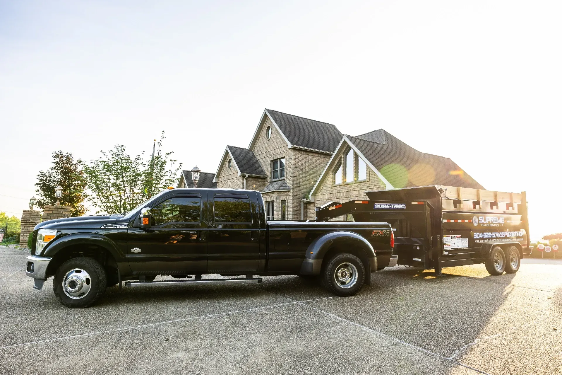 Black pickup truck with trailer, parked in front of a large house. The trailer appears to be carrying materials.