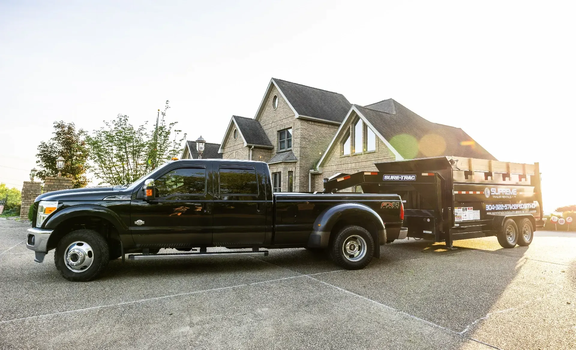 Black pickup truck towing a trailer loaded with wood, parked on gravel in front of a large house.