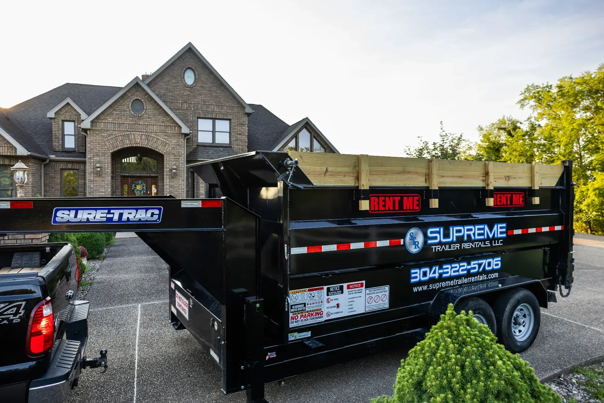 A black Sure-Trac dump trailer with Supreme Trailer Sales branding is parked in a driveway in front of a brick house.