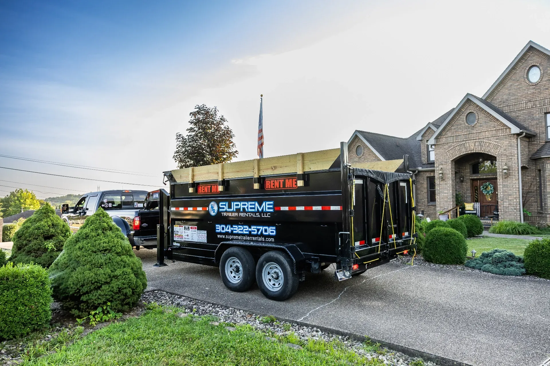 A black Supreme dump trailer hitched to a pickup truck parked in a driveway in front of a brick house.