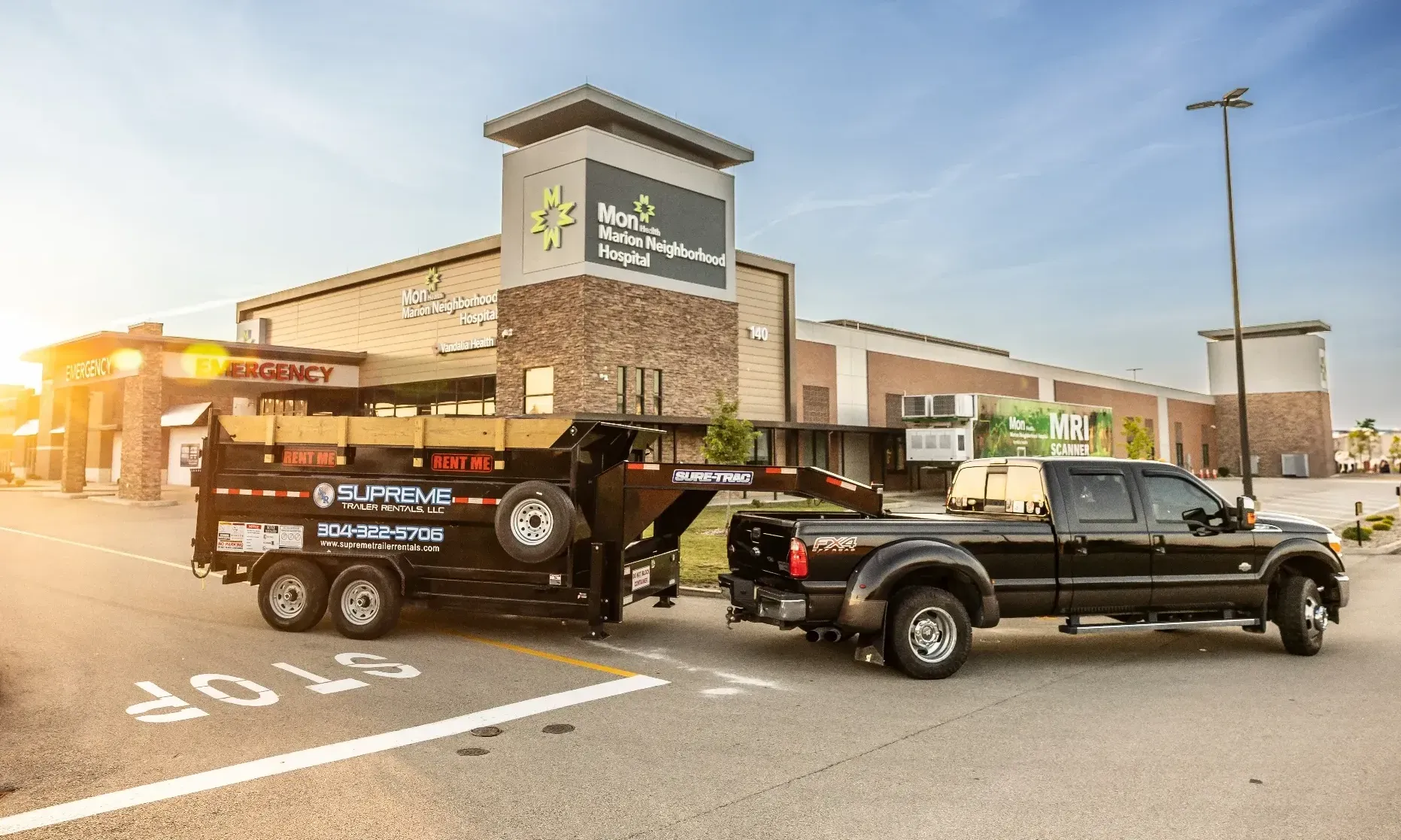 Black truck towing a trailer loaded with materials in front of a large store under a sunny sky.