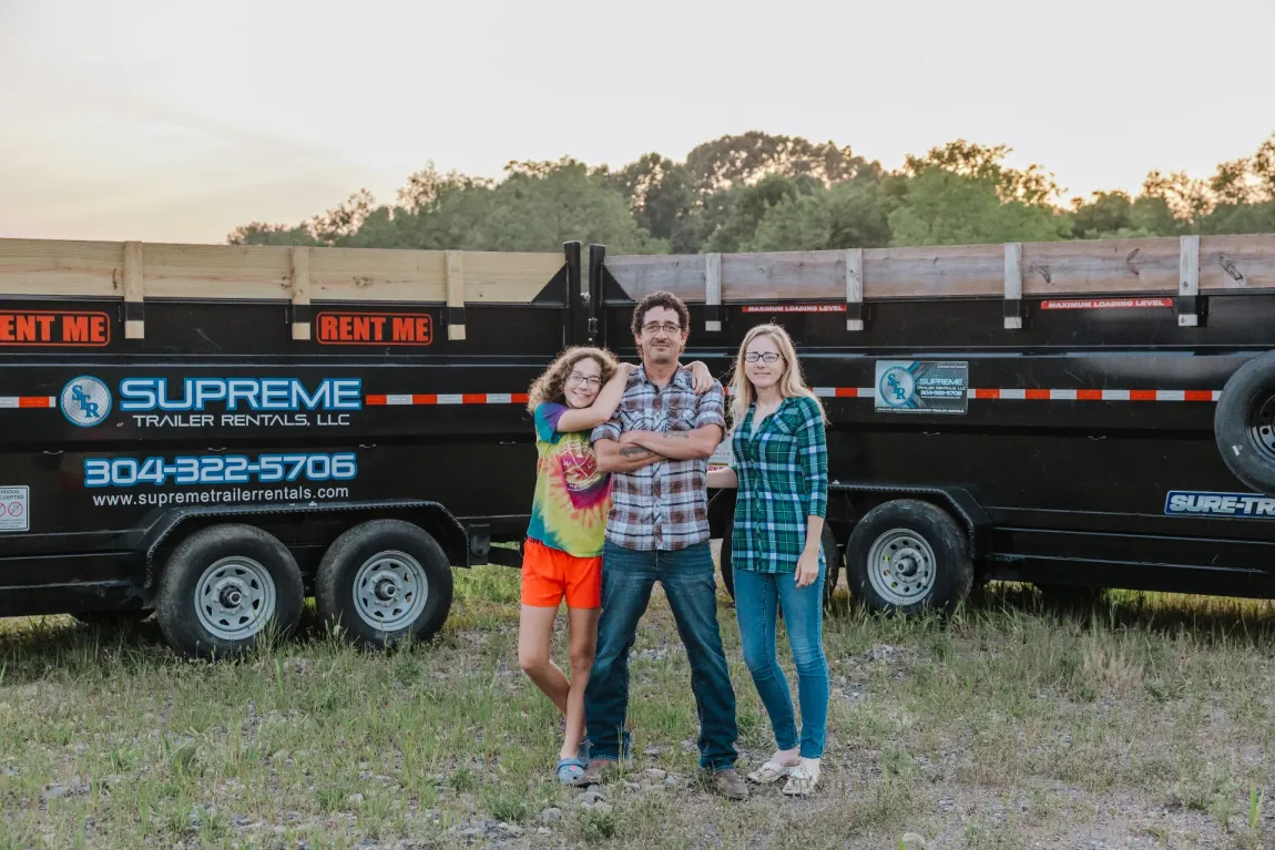 Three people stand in front of a black dump trailer. A man with arms crossed is flanked by two women. All three are smiling, outdoors.