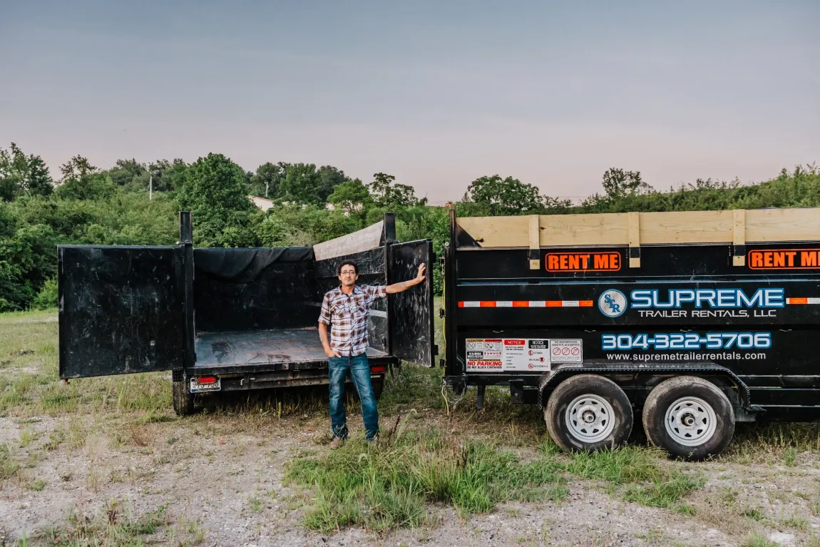 A man stands next to two black dump trailers in an outdoor setting. The trailers are labeled 