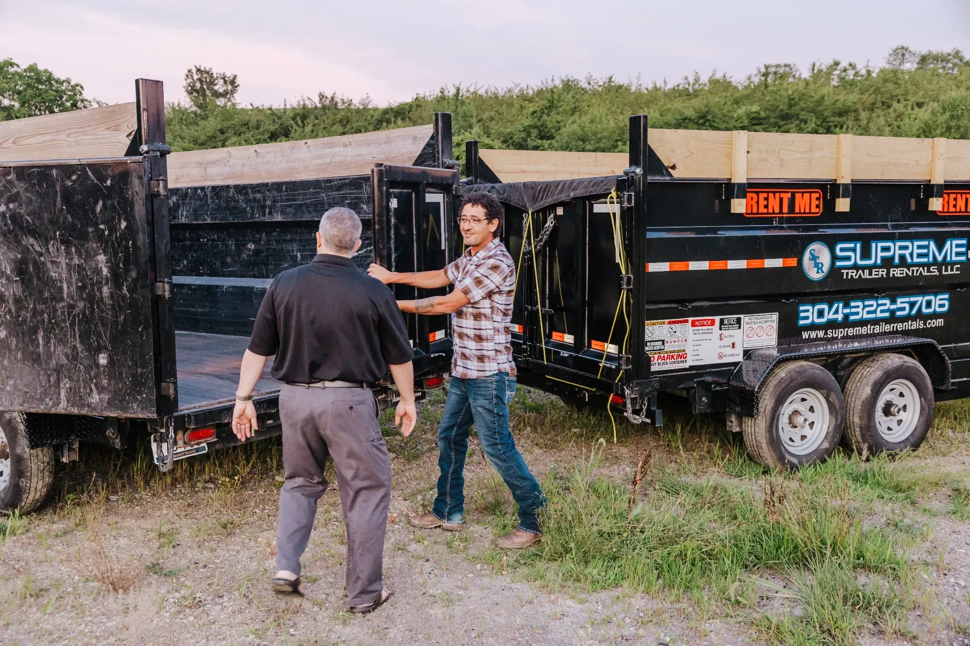 Two men stand near black dump trailers on grass. One opens a trailer door; the other, in a black shirt, faces him.