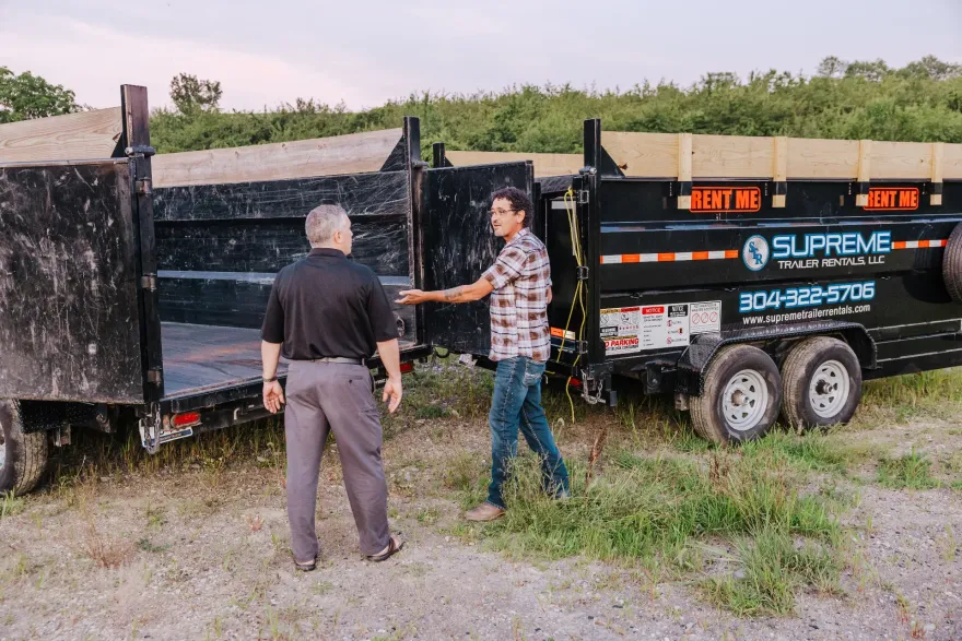 Two men near black Supreme Rentals dump trailers outdoors. One man points towards the trailer while the other faces him.