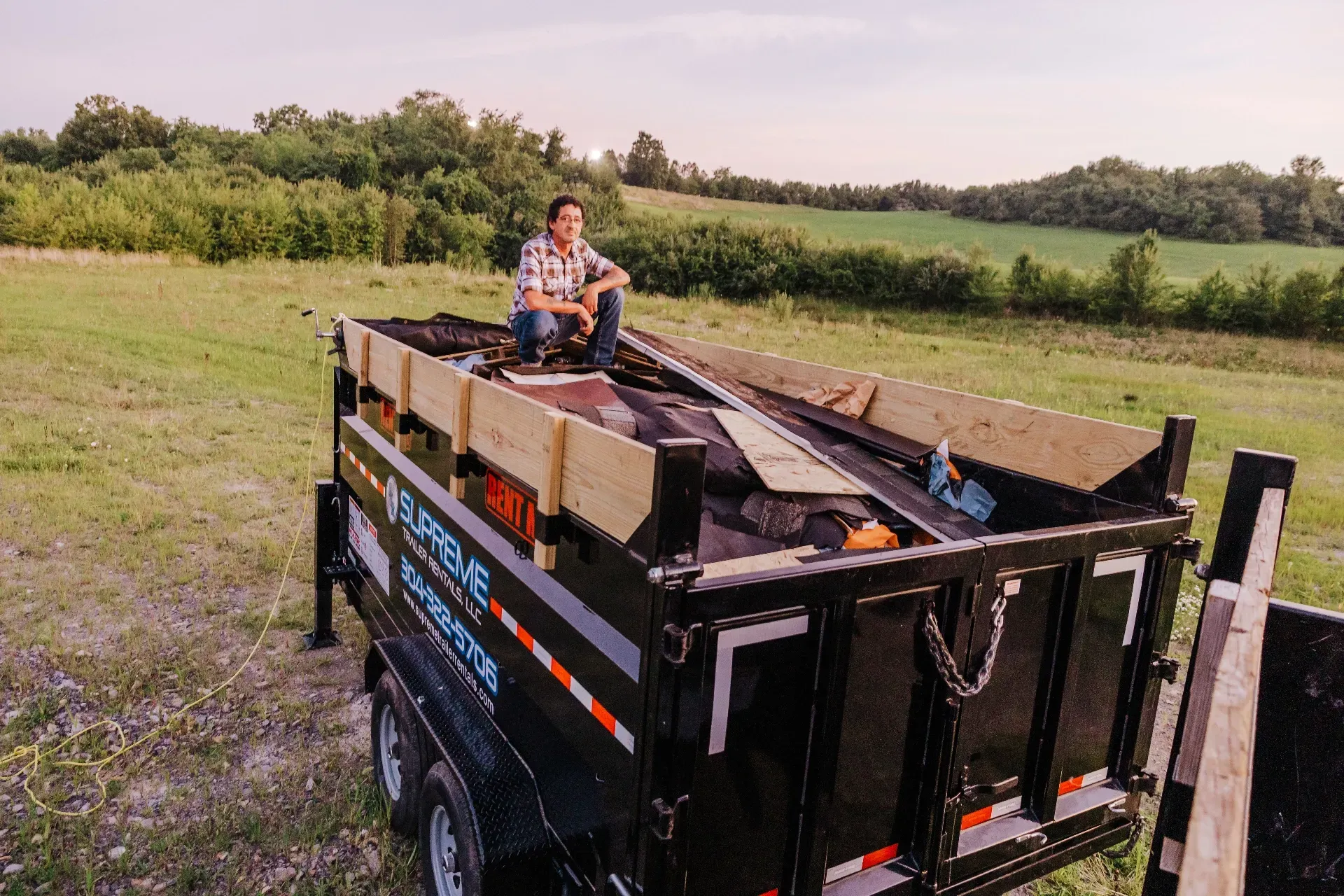 Person sits atop a loaded black trailer with wood planks, in a grassy field under a dusky sky, with a tree line in the background.