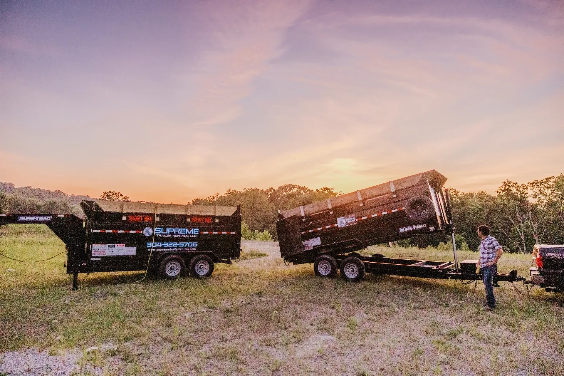 Two black dump trailers on grass field, one tilted to dump. A man stands by the truck, sunset sky background.