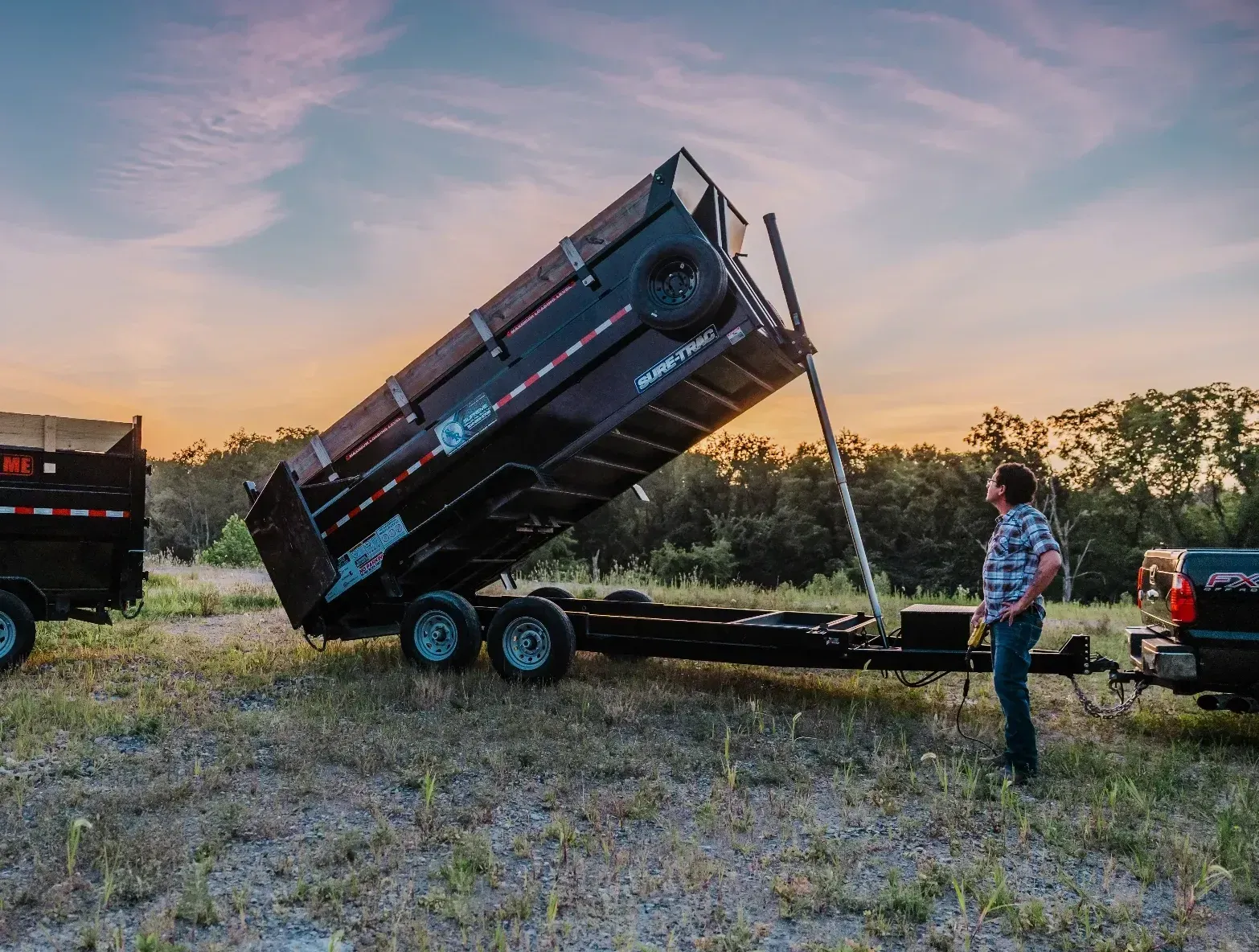 A man watches a black dump trailer tilting up, beside a pickup truck, with a sunset in the background.