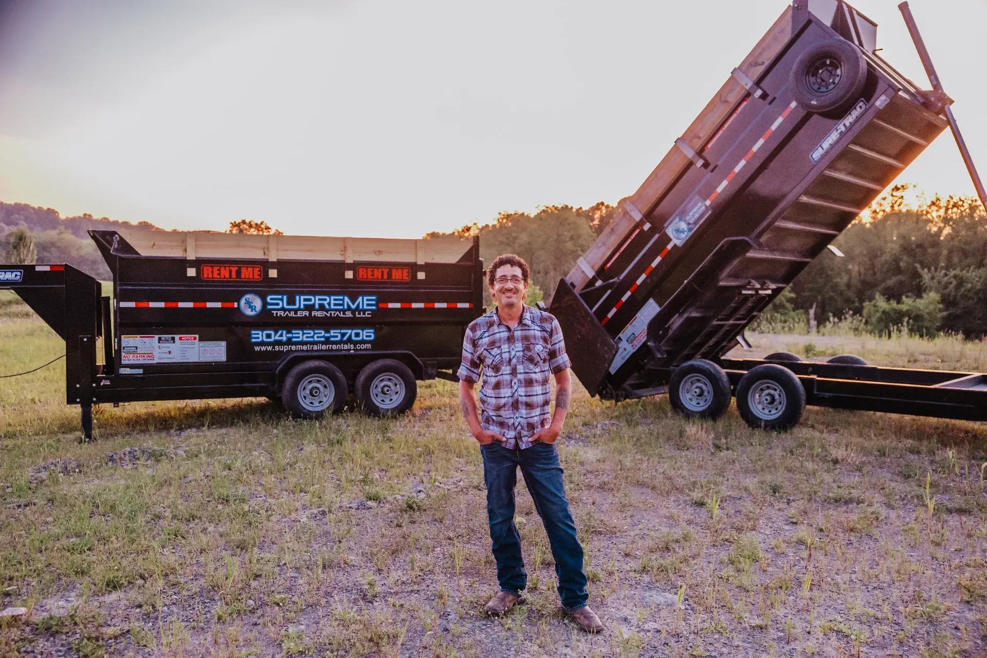 Man standing in a field next to a black dump trailer, with the bed tilted up. Sunset in the background.