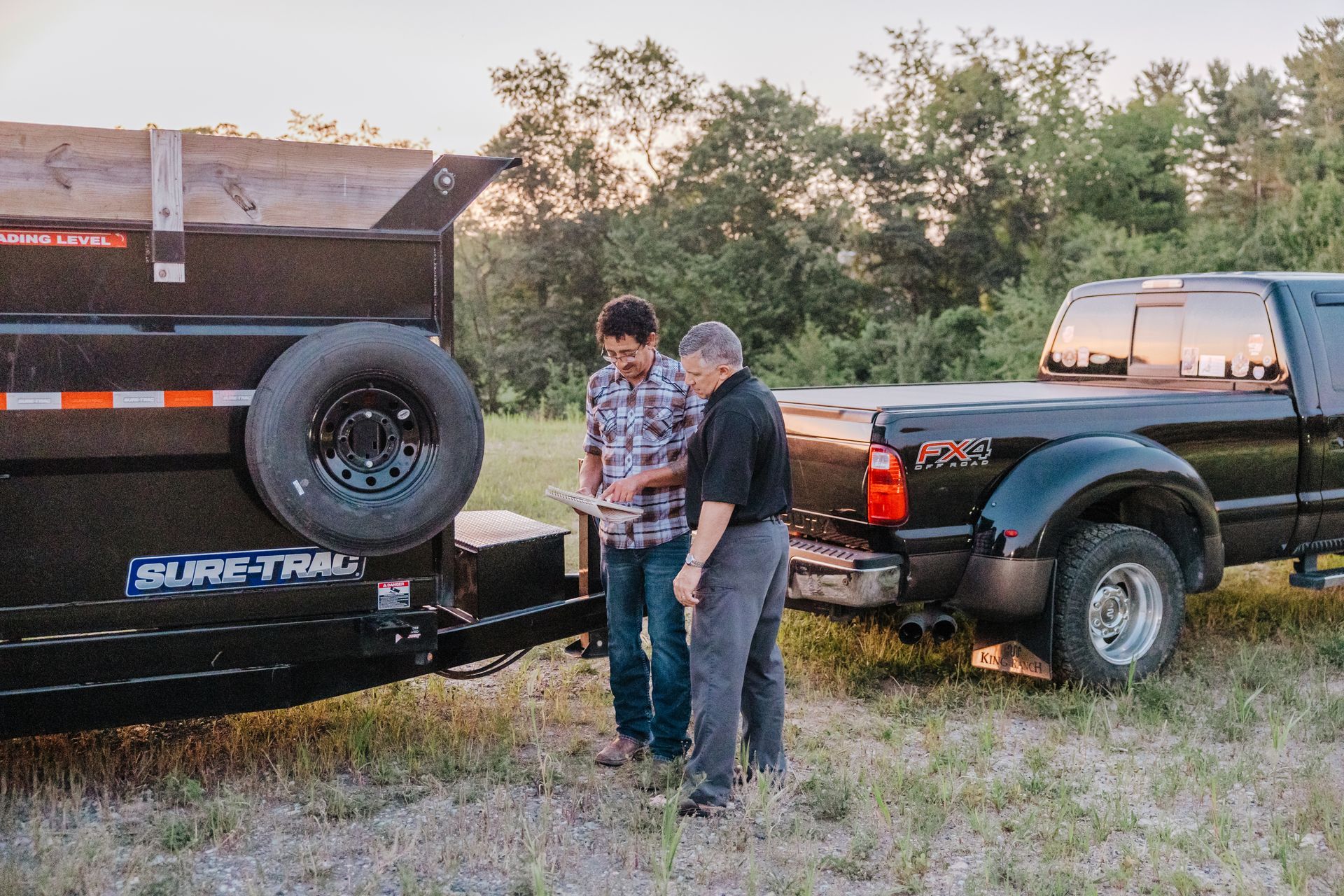 Two men standing near a black truck with a trailer. One man is looking at a phone, while the other looks on, in a field.