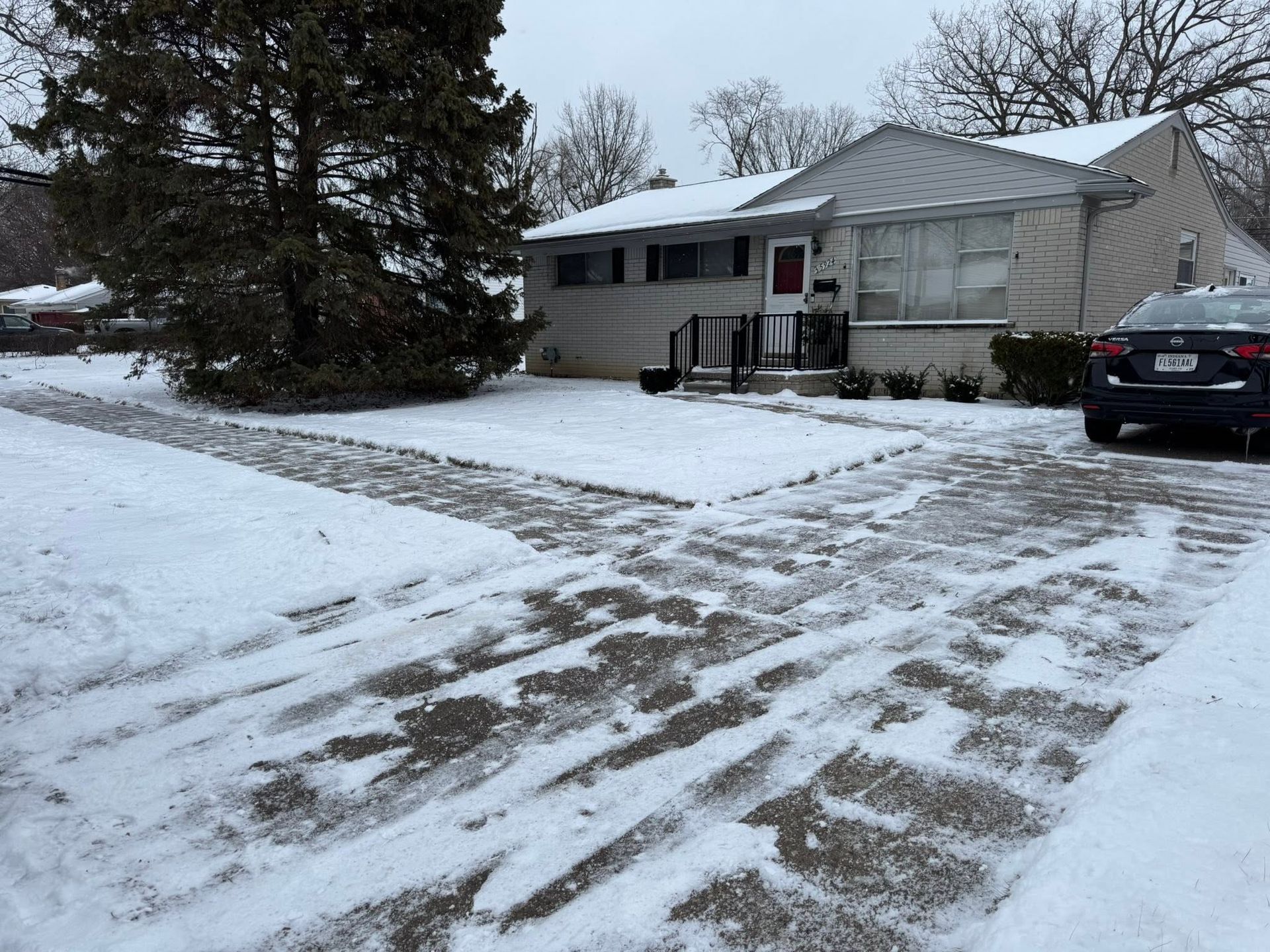 Snow-covered suburban house with driveway, a parked car, and a large tree in the front yard.
