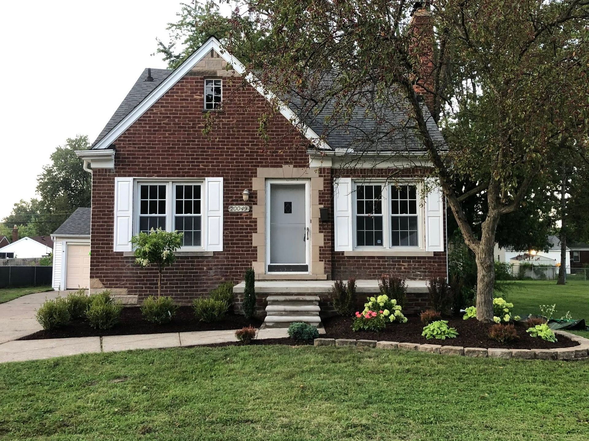Small brick house with white trim, front steps, two windows, and a landscaped yard with trees and flowers