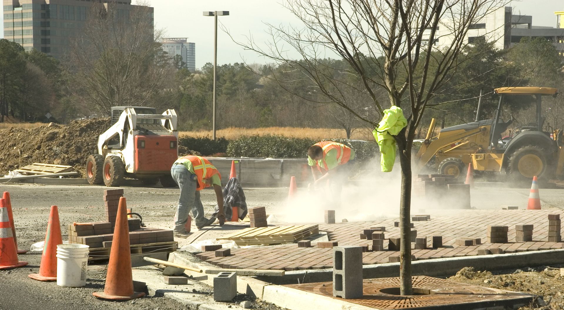 Construction workers repair a road, with orange cones and concrete dust at a work site.