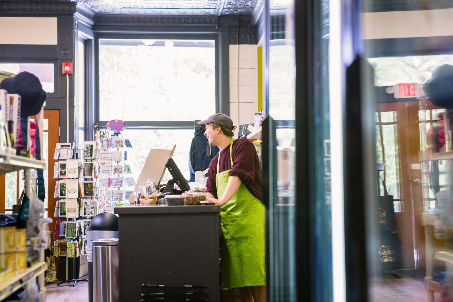 Barista in green apron serving at a coffee shop counter with a bright window behind.
