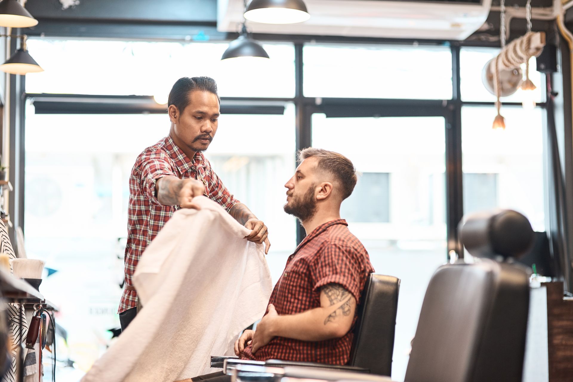 Stylist drapes a white cape over a seated client in a bright barbershop.