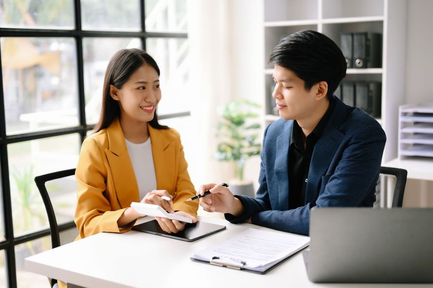 Two coworkers discuss notes at a bright office table with a laptop and notebook.