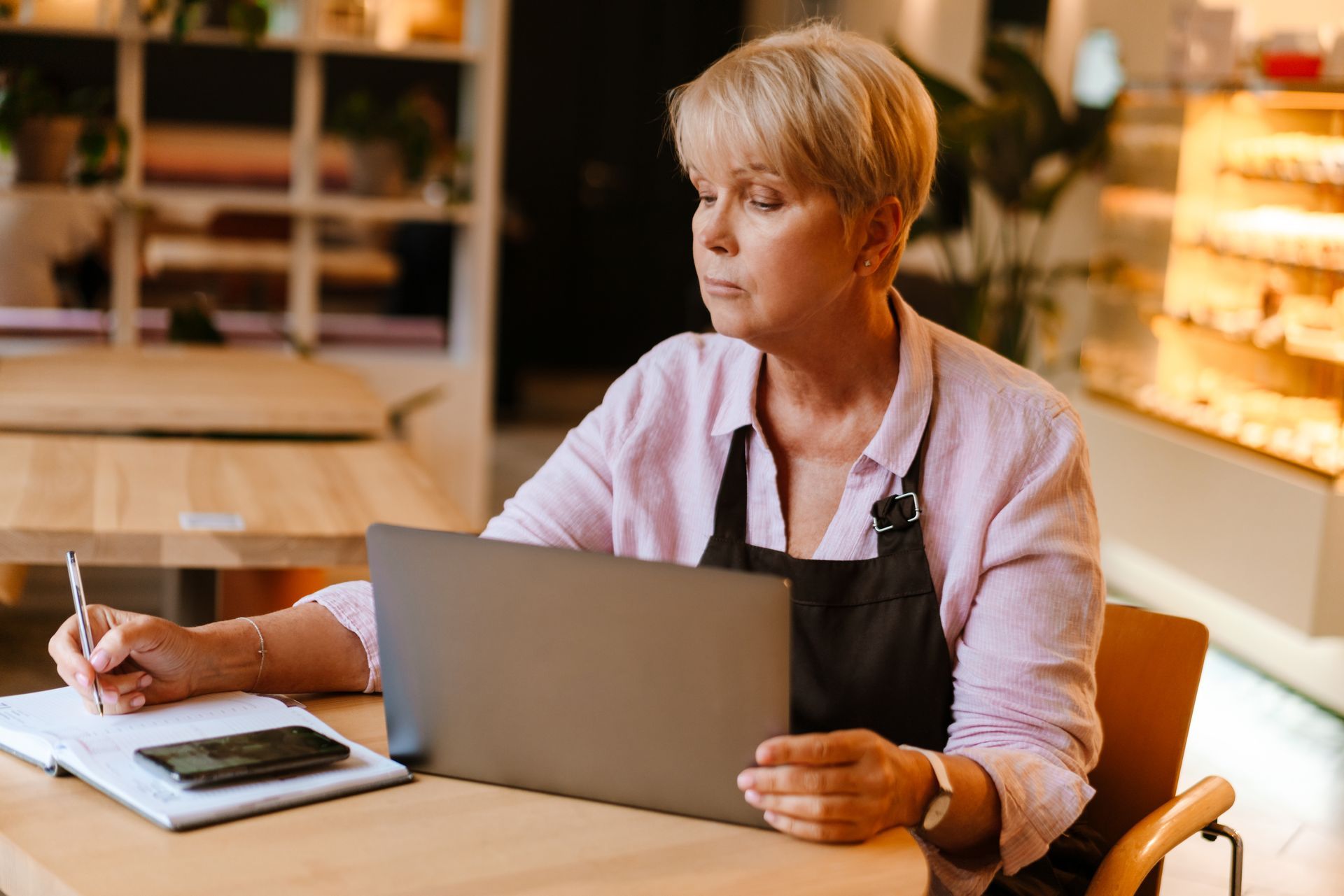 Person working on a laptop at a café table, reading a notebook with a warm, blurred background