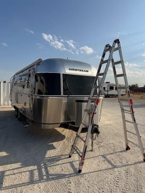 Silver Airstream trailer in a gravel lot with two ladders leaning nearby under a blue sky