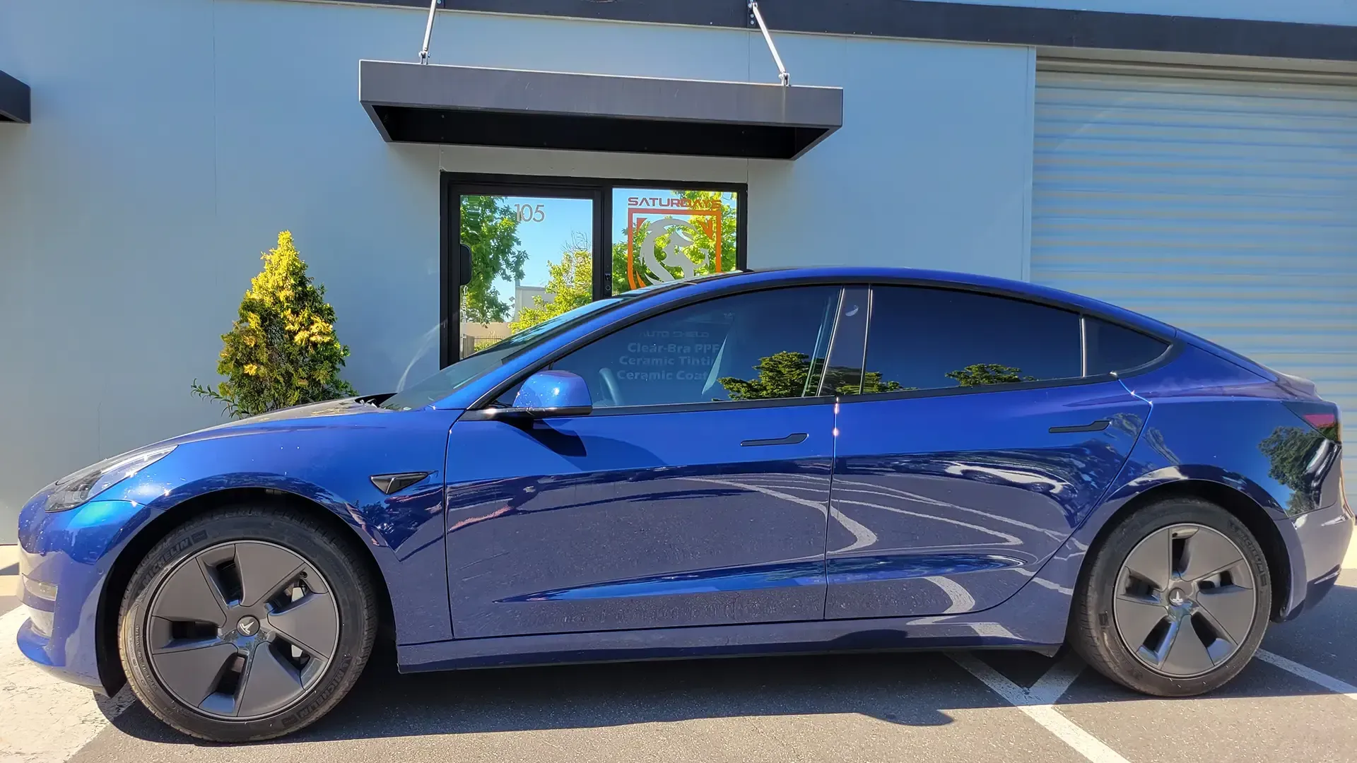 A blue tesla model 3 is parked in a parking lot in front of a building.