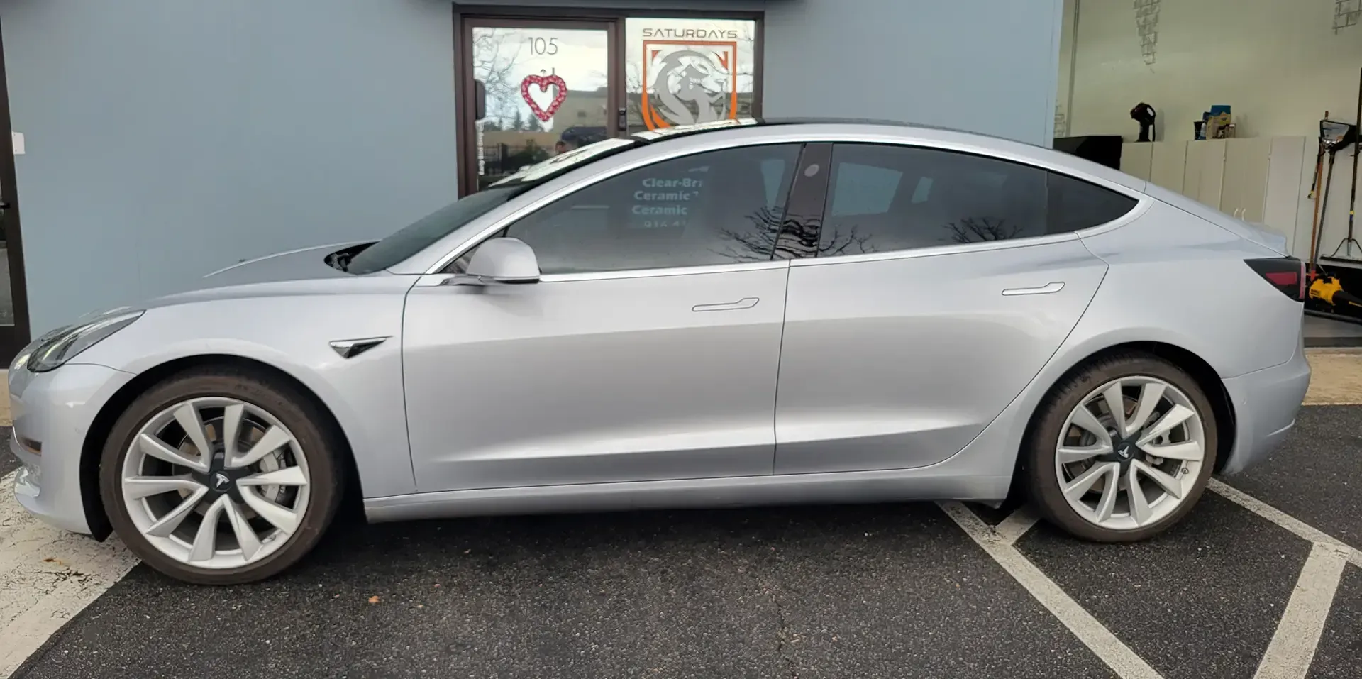 A silver tesla model 3 is parked in a parking lot in front of a building.