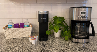 Kitchen counter with coffee maker, water dispenser, potted plant, and basket of drinks against a tiled backsplash